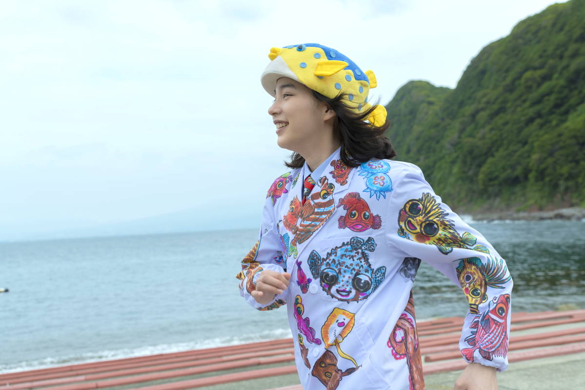 のん　頭にハコフグ帽子、たくさんの色鮮やかな魚の絵が描かれた服で笑顔　「さかなのこ」場面写真